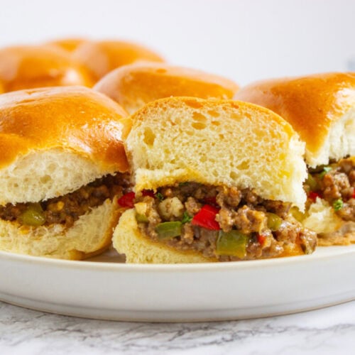 Three sloppy joe sliders with ground beef, green and red peppers, and onions, served on a white plate with more sliders in the background.