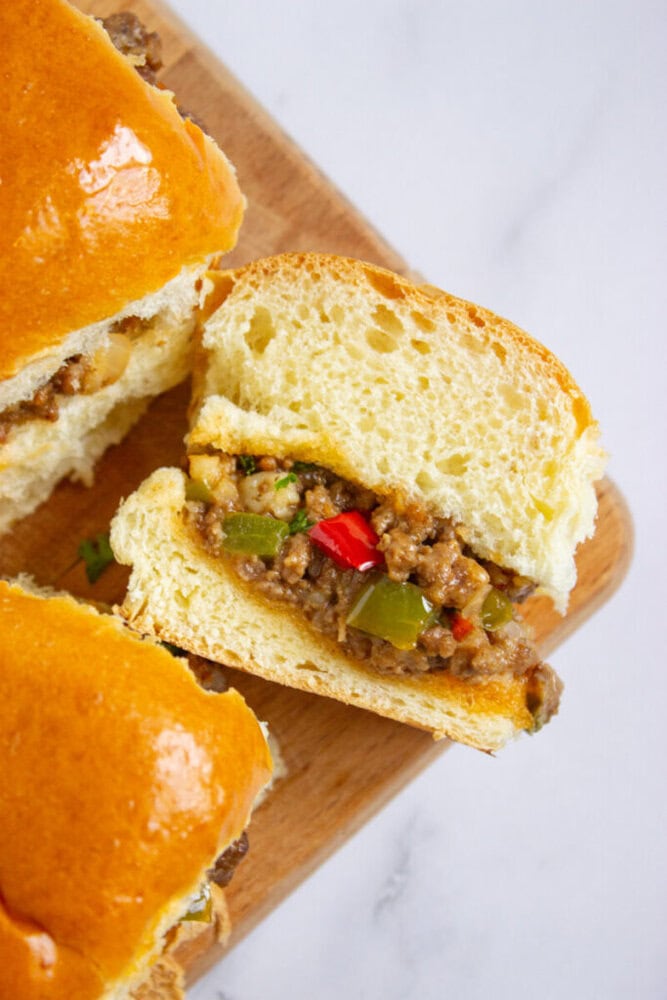 A close-up of a sandwich with ground beef, chopped bell peppers, and onions on a wooden board, featuring soft, glossy bread.