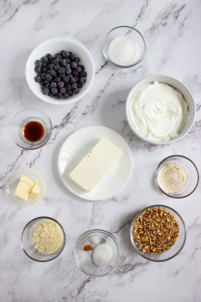 Various cheesecake ingredients, including blueberries, sugar, cream cheese, butter, vanilla extract, flour, almond flour, and chopped nuts, arranged in bowls on a marble surface.