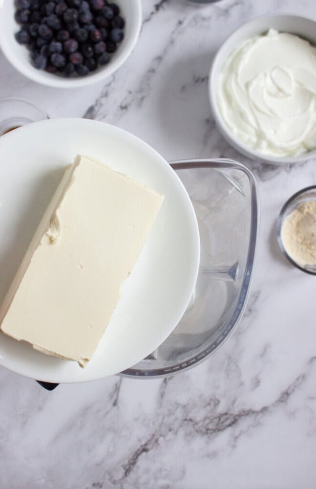 A block of cream cheese on a white plate is held over a blender surrounded by bowls of blueberries, Greek yogurt, and protein powder on a marble countertop.