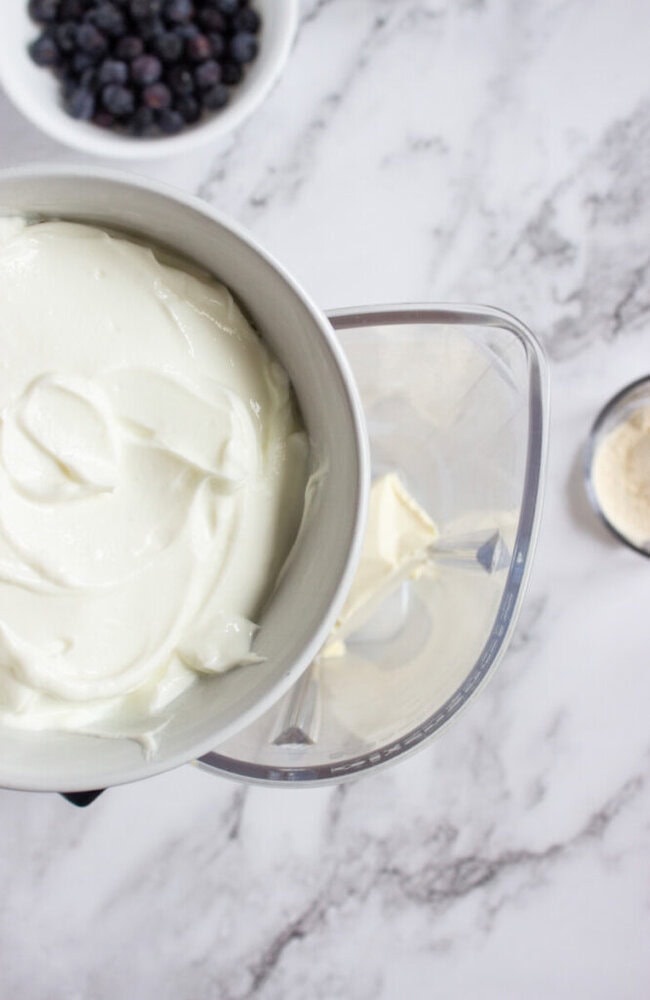 A bowl of yogurt is being poured into a blender, with a bowl of blueberries and a small container of powder nearby on a marble countertop.