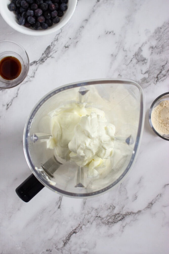 A blender with yogurt inside sits on a marble counter, surrounded by bowls containing blueberries, vanilla extract, and protein powder.