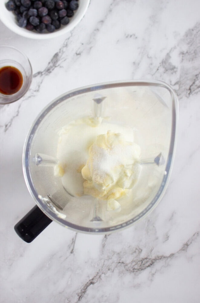 A blender contains yogurt and sugar on a marble countertop, with a bowl of blueberries and a glass of liquid nearby.