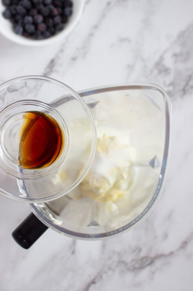 A hand pours vanilla extract from a glass cup into a blender containing cream cheese, sugar, and yogurt on a marble countertop; blueberries are in a bowl nearby.