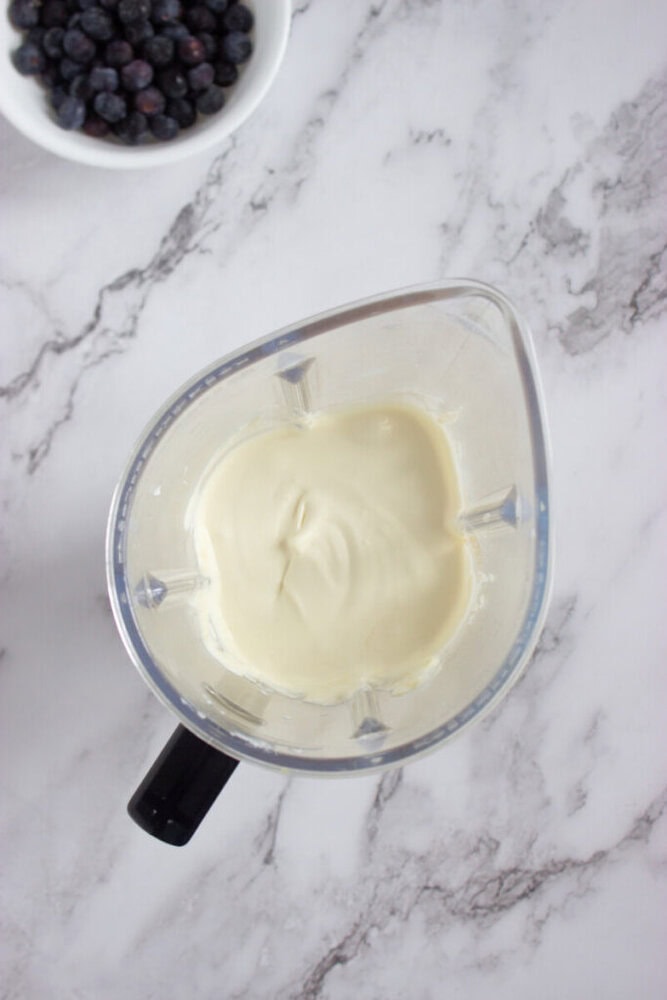 A blender cup filled with a thick, creamy white mixture sits on a marble countertop; a bowl of blueberries is nearby in the upper left corner.