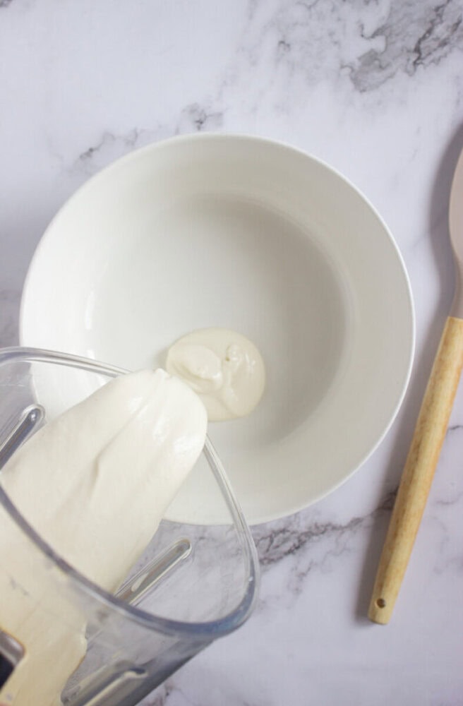 A blender is pouring a thick white creamy mixture into an empty white bowl on a marble surface. A wooden spatula lies beside the bowl.