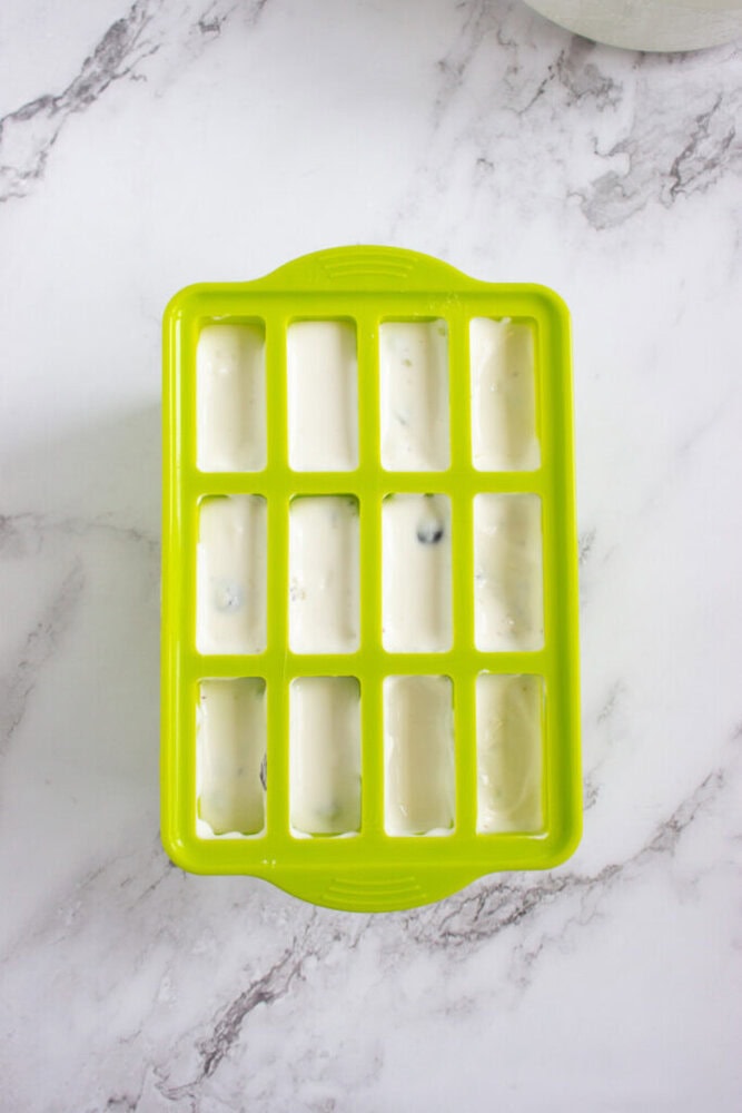 A green silicone ice cube tray filled with a white creamy mixture sits on a marble countertop.