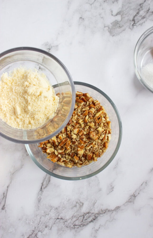 Three glass bowls on a marble surface, one with almond flour, one with chopped nuts, and a partially visible bowl with granulated sugar.