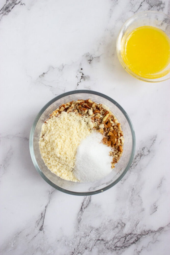 A glass bowl with almond flour, granulated sugar, and brown sugar on a marble surface; a separate small bowl contains melted butter.