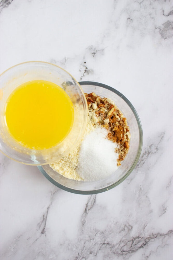 A glass bowl with dry ingredients including sugar and crumbs, with melted butter being poured in from another bowl, on a marble surface.