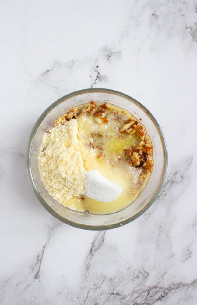 A glass bowl containing chopped nuts, almond flour, granulated sugar, and melted butter on a white marble surface.