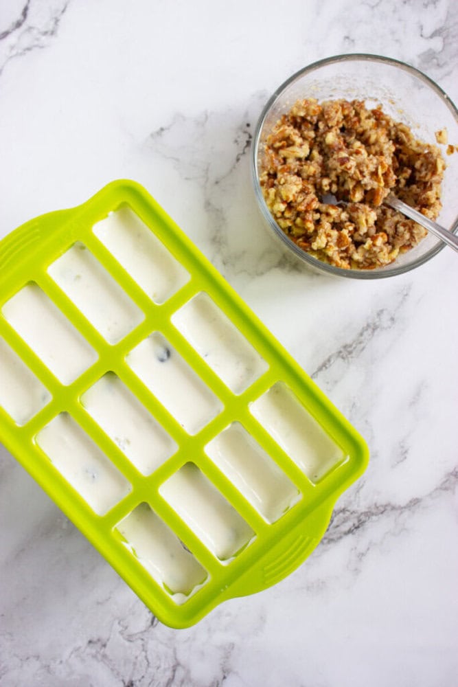 A green silicone ice cube tray filled with white liquid sits next to a glass bowl of mixed ingredients with a spoon on a marble surface.