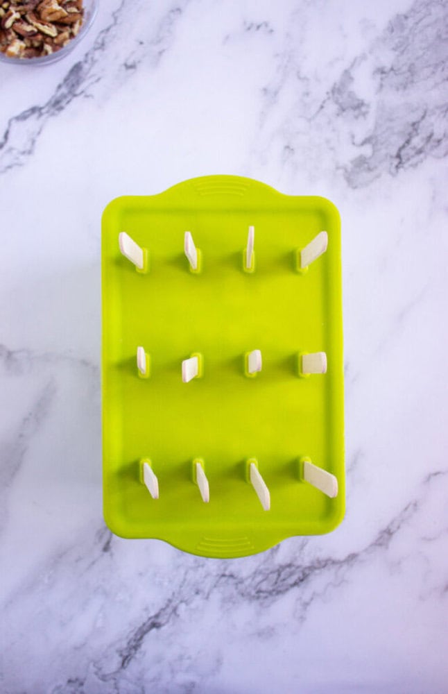 A green popsicle mold with wooden sticks inserted, placed on a white marble surface.