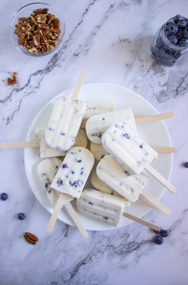 A plate of blueberry yogurt popsicles with visible blueberries is arranged on a marble surface, surrounded by a jar of blueberries and a bowl of chopped pecans.