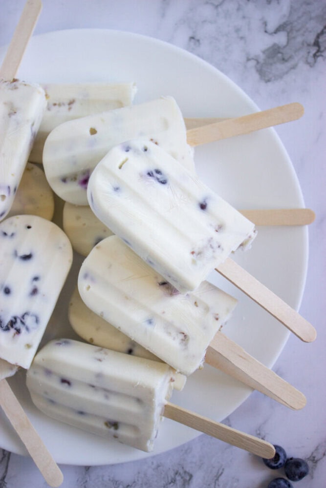 A plate of creamy white popsicles with visible bits of blueberries, arranged on a white marble surface.