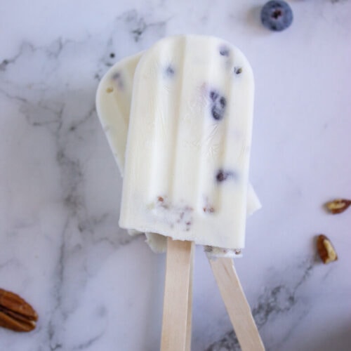 Two yogurt popsicles with blueberries and nuts on a marble surface, surrounded by loose blueberries and pecans.