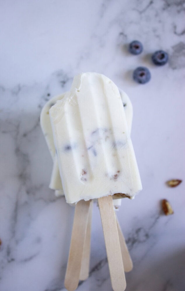Two yogurt and blueberry popsicles on a marble surface, with one having a bite taken out of it. Blueberries and small nut pieces are scattered nearby.