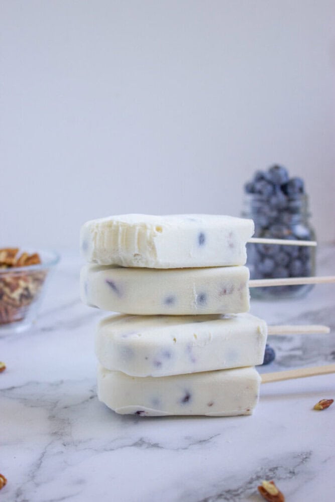 Four blueberry yogurt popsicles are stacked on a marble surface, with a bite taken out of the top one. Bowls of blueberries and pecans are in the background.
