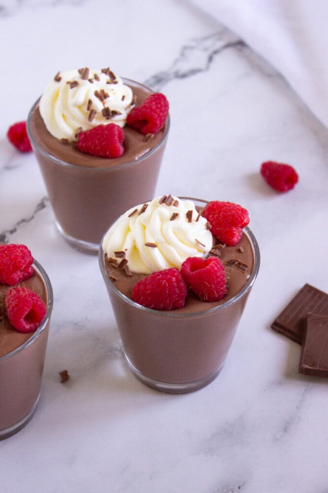 Three glasses of chocolate mousse topped with whipped cream, fresh raspberries, and chocolate shavings on a marble surface.