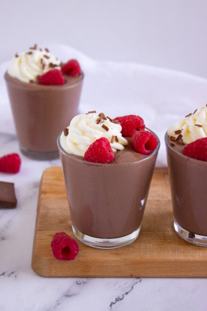 Three glasses of chocolate mousse topped with whipped cream, chocolate shavings, and fresh raspberries, placed on a wooden board with extra raspberries nearby.