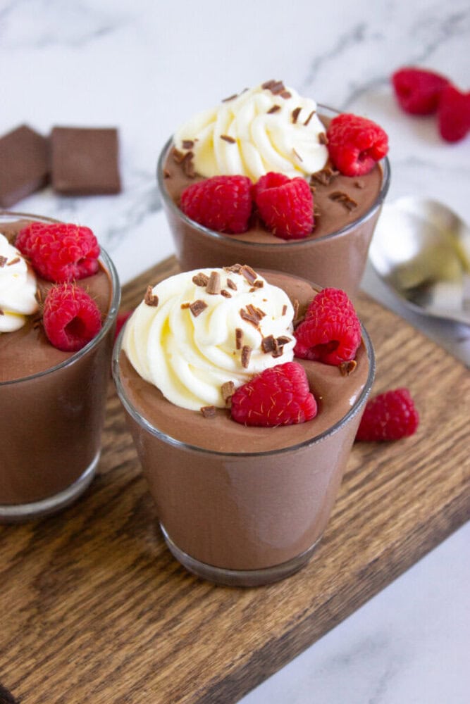 Three glasses of chocolate mousse topped with whipped cream, raspberries, and chocolate shavings are displayed on a wooden board, with chocolate pieces and a spoon in the background.