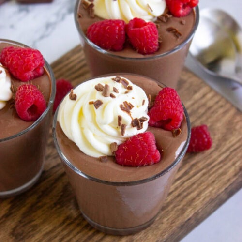 Three glasses of chocolate mousse are topped with whipped cream, chocolate shavings, and fresh raspberries, displayed on a wooden board with a spoon nearby.