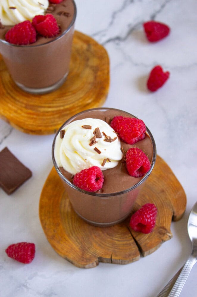A glass of chocolate mousse topped with whipped cream, chocolate shavings, and raspberries, set on a wooden coaster with extra raspberries nearby.