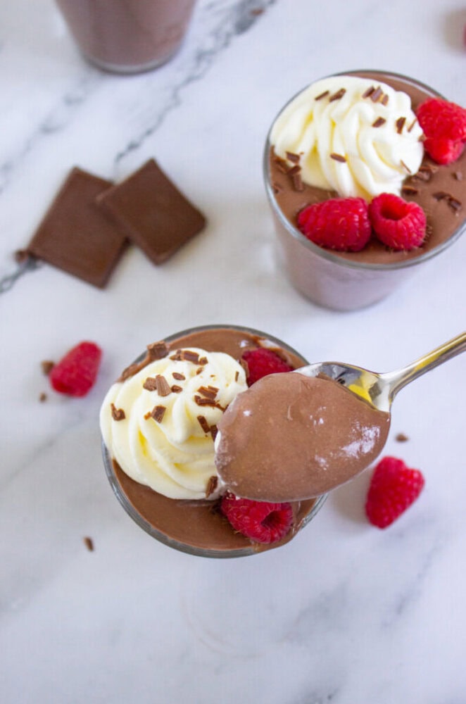 A spoon holding chocolate mousse above a glass topped with whipped cream, raspberries, and chocolate shavings; chocolate squares and raspberries nearby.