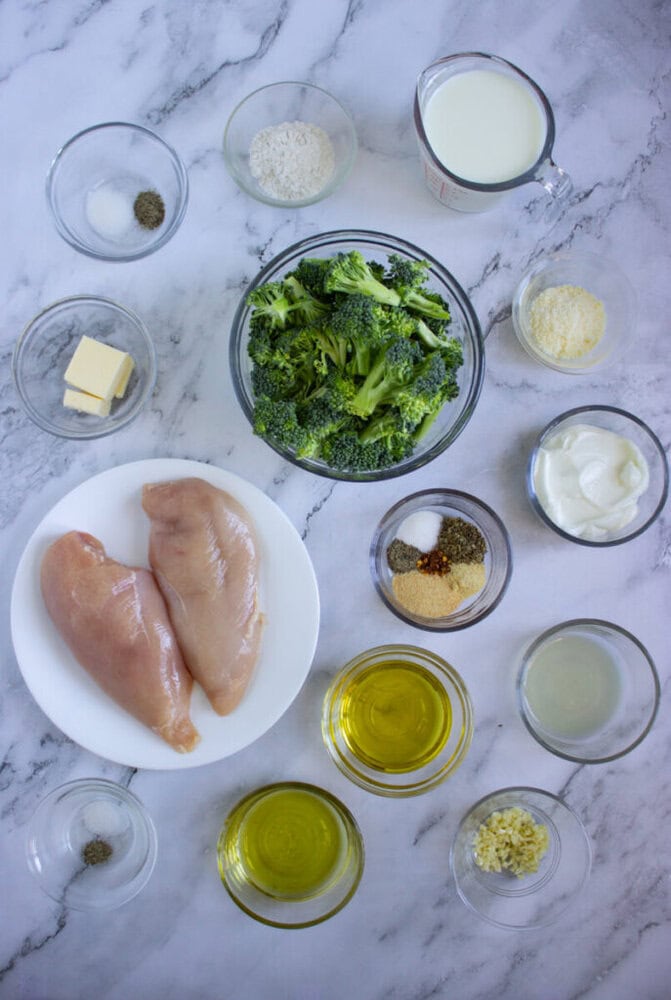 Ingredients for a chicken and broccoli dish, including raw chicken breasts, broccoli, olive oil, butter, spices, garlic, flour, milk, parmesan, sour cream, and lemon juice, arranged on a marble surface.