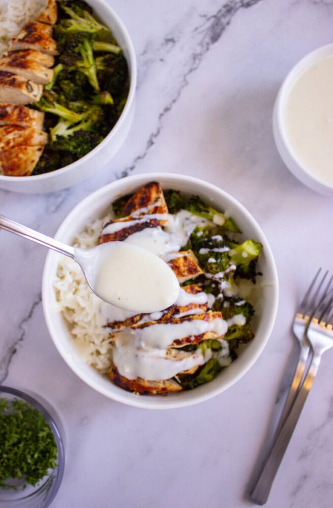 A spoon drizzles white sauce over a bowl of rice, grilled chicken, and broccoli. Nearby are two forks, a bowl of sauce, and a small dish of chopped herbs on a marble surface.