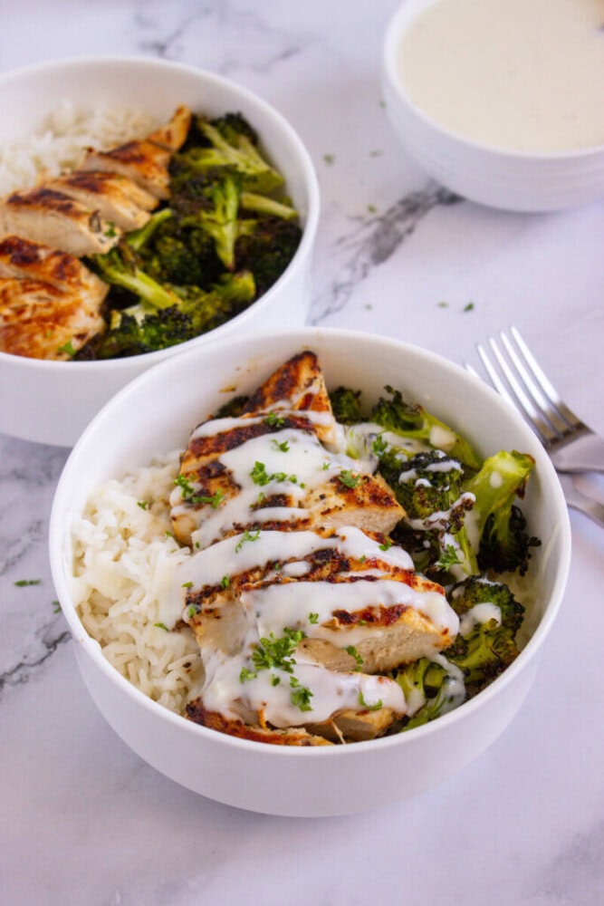 Two white bowls filled with rice, grilled chicken, and broccoli, topped with a creamy white sauce. A bowl of extra sauce and a fork are set on a marble surface nearby.