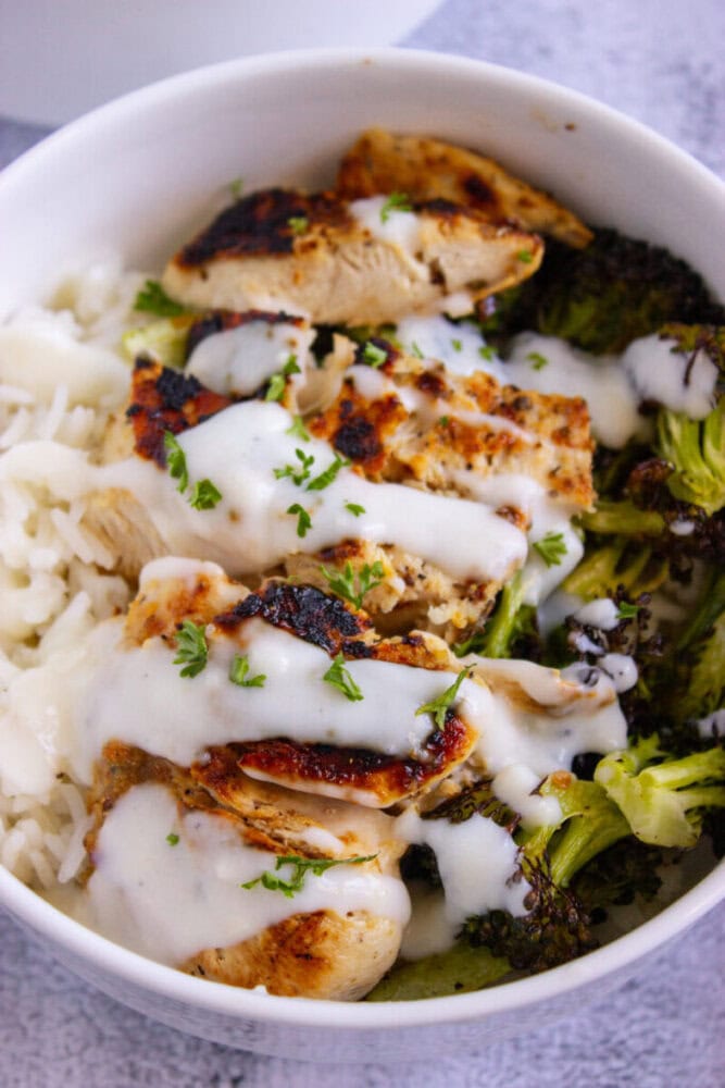 A bowl of white rice topped with grilled chicken slices, roasted broccoli, and drizzled with white sauce, garnished with chopped herbs.