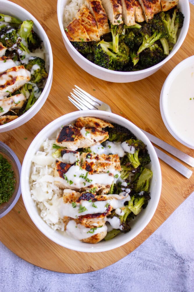 Three bowls of grilled chicken, broccoli, and rice topped with white sauce on a round wooden board, with a fork, sauce bowl, and chopped herbs on the side.