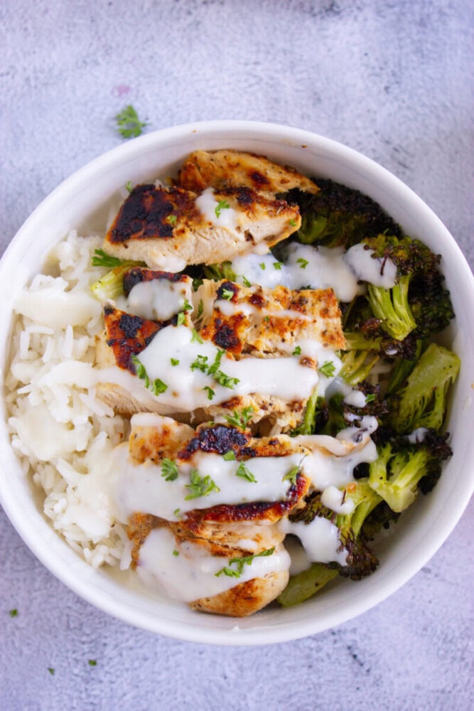 A bowl of white rice topped with grilled chicken slices, roasted broccoli, and a drizzle of white sauce, garnished with chopped herbs.