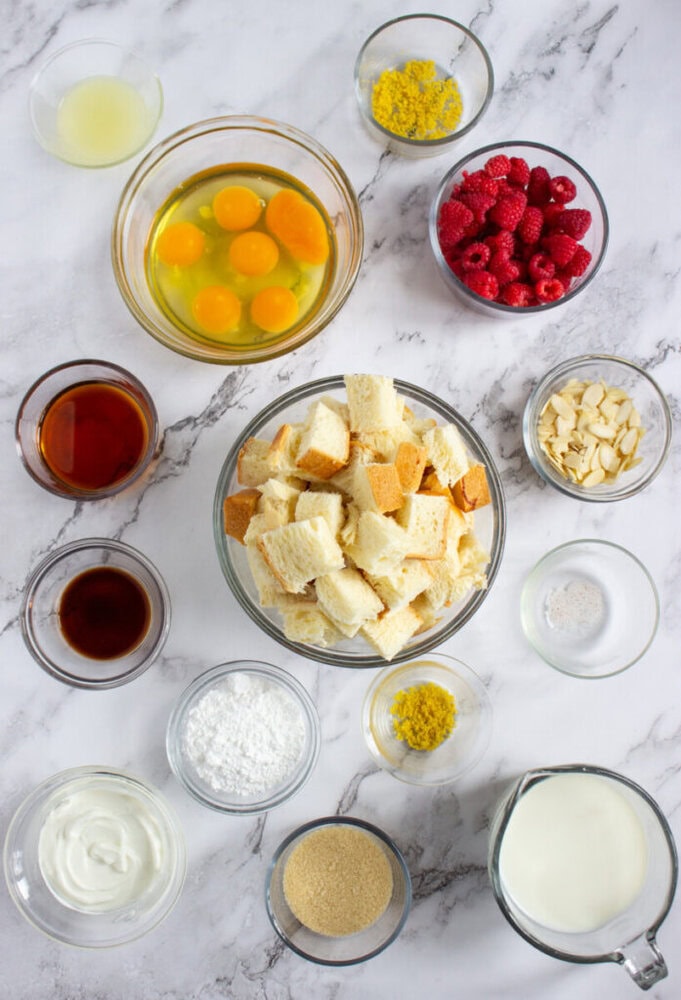 A variety of ingredients in glass bowls on a marble surface, including eggs, bread cubes, raspberries, milk, sugar, yogurt, vanilla, lemon zest, almonds, and powdered sugar.