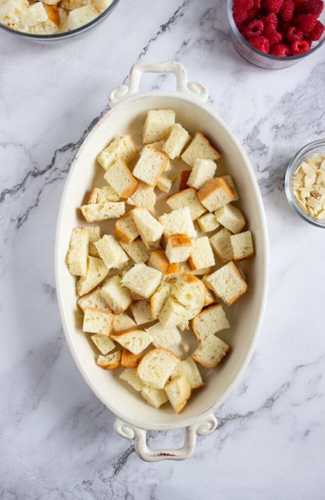 An oval baking dish filled with cubed bread sits on a marble countertop, with bowls of raspberries and sliced almonds nearby.