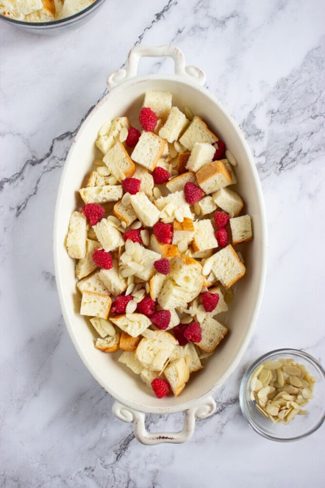 An oval baking dish filled with cubed bread, raspberries, and sliced almonds on a marble surface. A small bowl of sliced almonds is nearby.