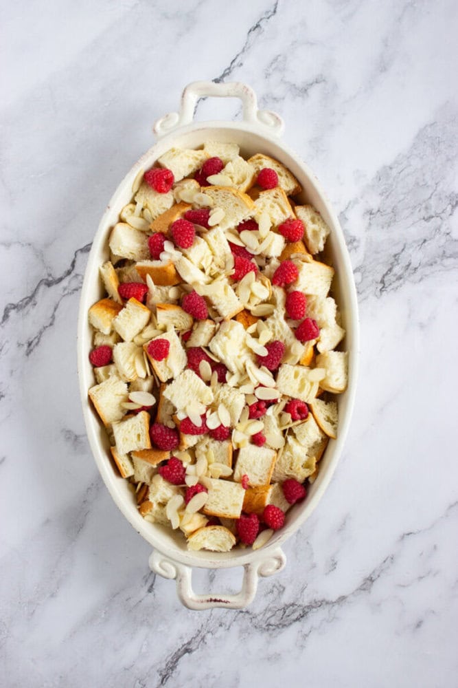 White oval baking dish filled with cubed bread, fresh raspberries, and sliced almonds on a marble countertop.