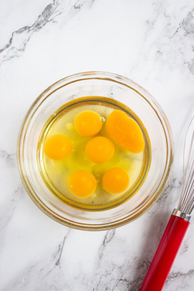 A glass bowl containing six raw eggs, cracked open with visible yolks, sits on a white marble surface next to a metal whisk with a red handle.