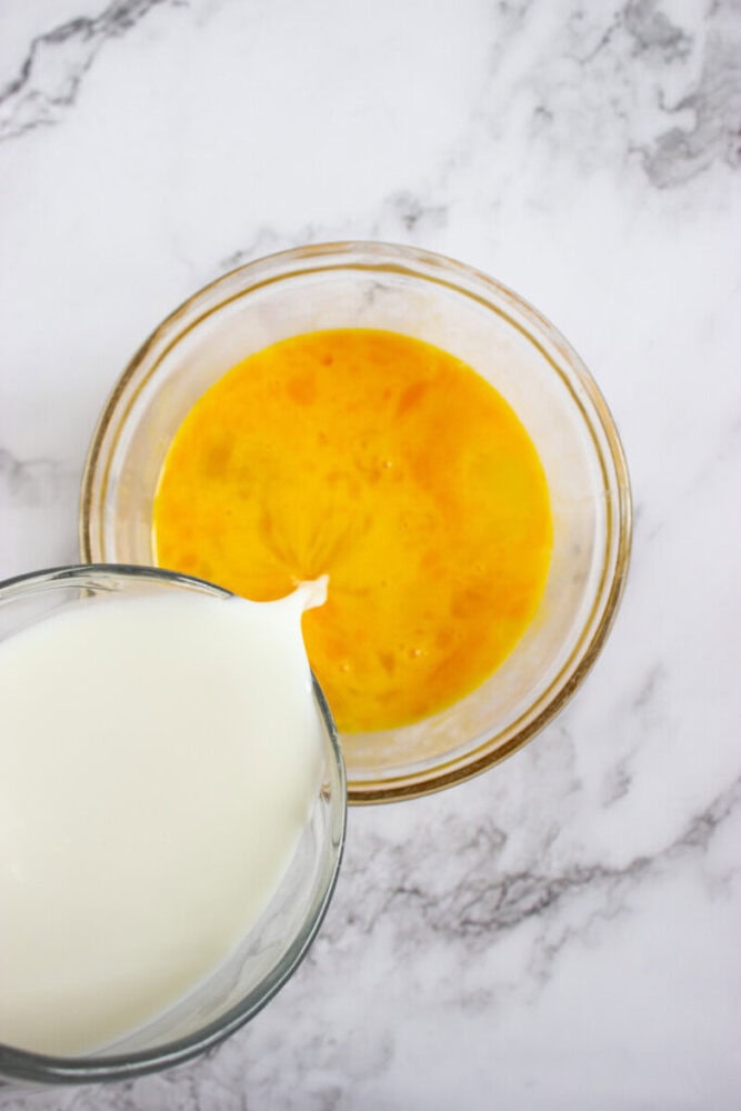 A glass bowl with beaten eggs sits on a marble surface while milk is being poured into it from a glass measuring cup.