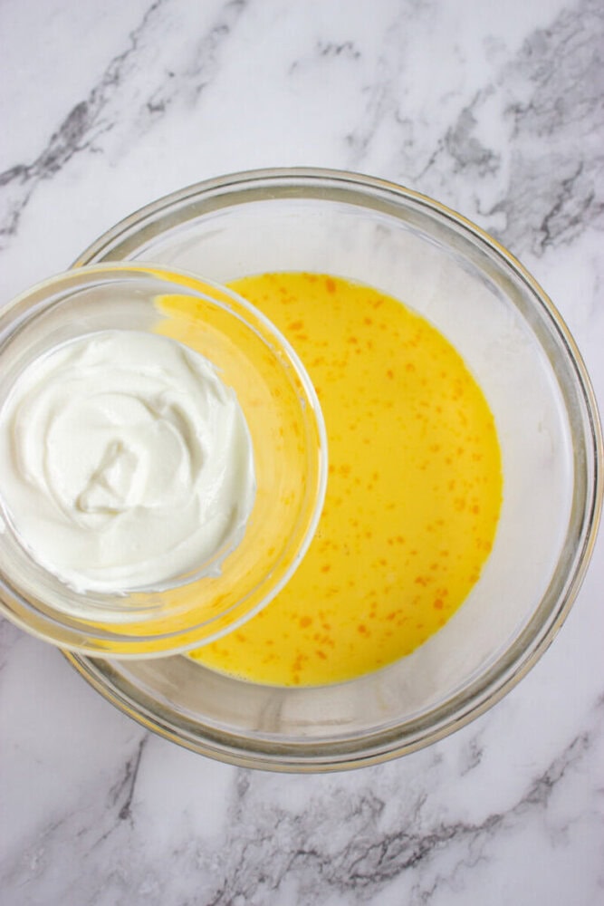 A glass bowl with a yellow liquid mixture next to a smaller bowl containing thick white yogurt, both on a marble countertop.
