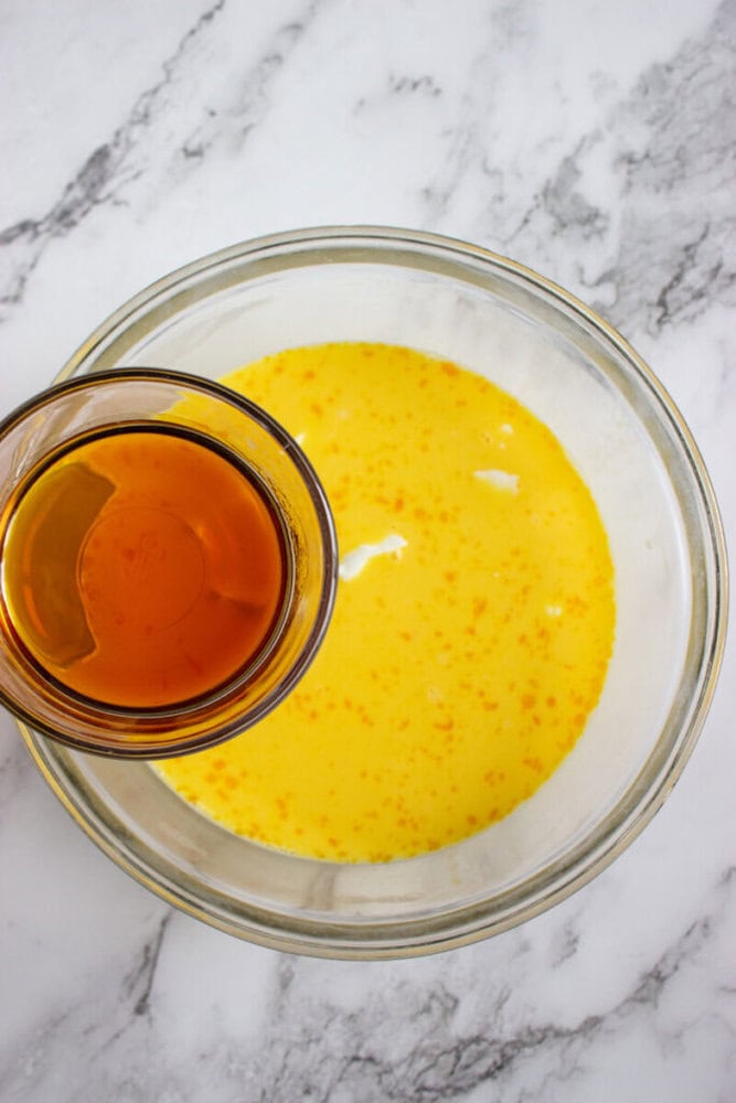 A glass bowl with a yellow liquid mixture, possibly eggs and milk, on a marble surface. A smaller glass container with a brown liquid is being held above the bowl.