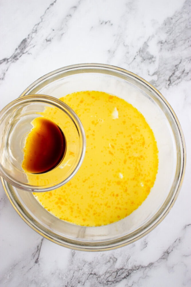 A small glass bowl with vanilla extract is being poured into a larger bowl containing a yellow liquid mixture on a marble surface.