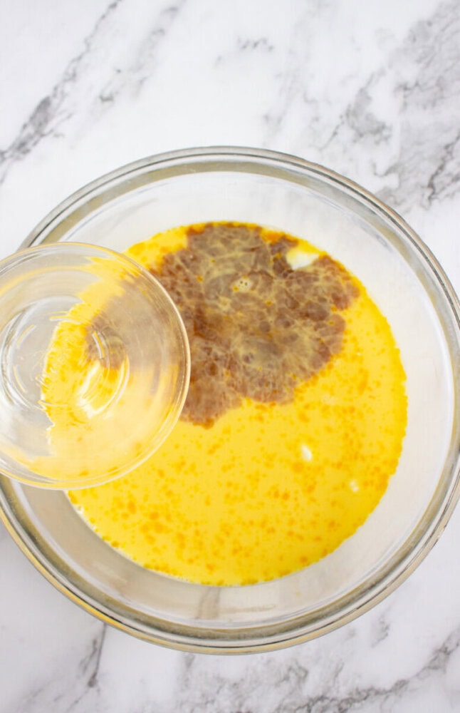A glass bowl containing a mixture of eggs and liquid with a hand pouring more liquid into it, set on a marble countertop.