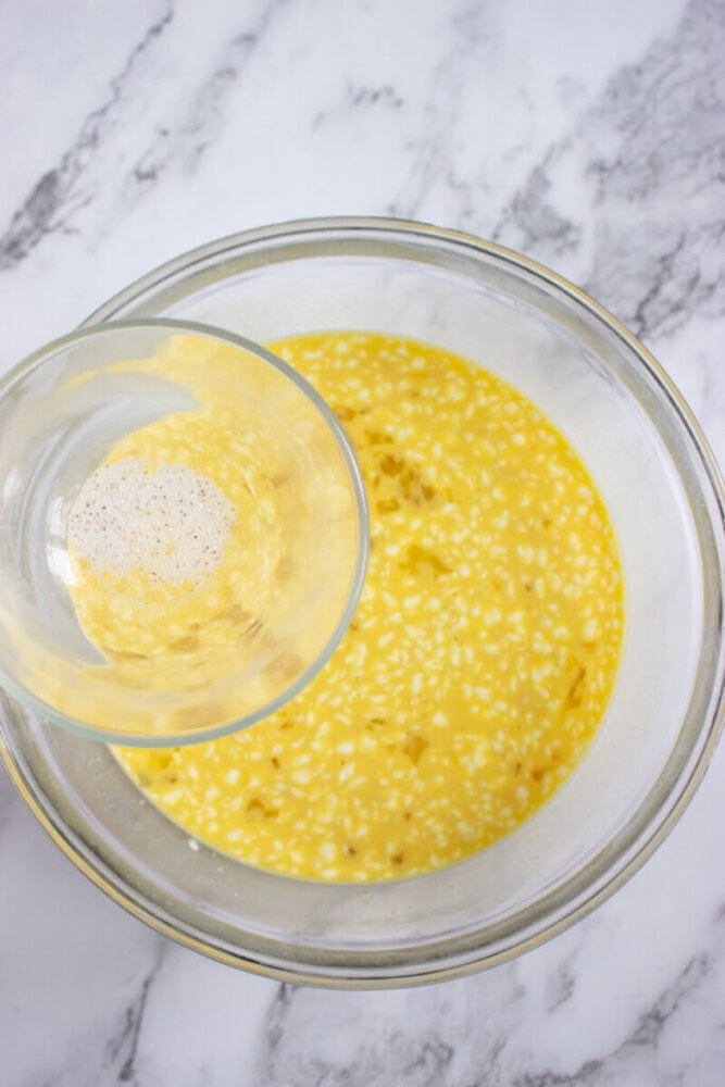 A glass bowl with a yellow egg mixture sits on a marble surface. Another small bowl with a liquid and spices is being poured into it.