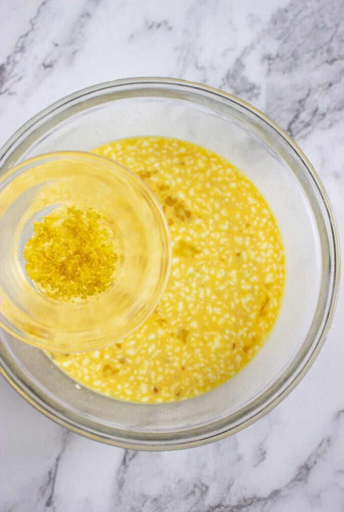 A glass bowl of yellow liquid mixture on a marble surface, with lemon zest being added from a smaller bowl above.