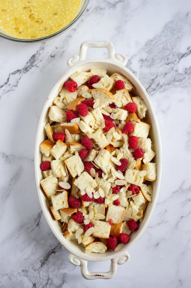 An oval baking dish filled with cubed bread, raspberries, and sliced almonds sits on a marble surface near a bowl of yellow liquid mixture.