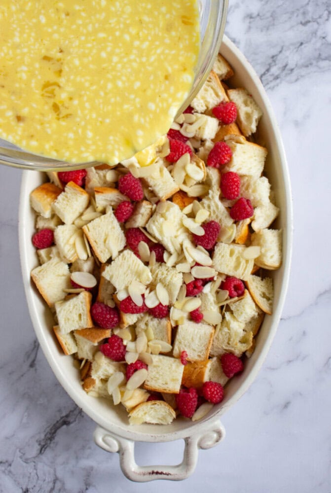 A baking dish with cubed bread, raspberries, and sliced almonds, with egg mixture being poured over the top.
