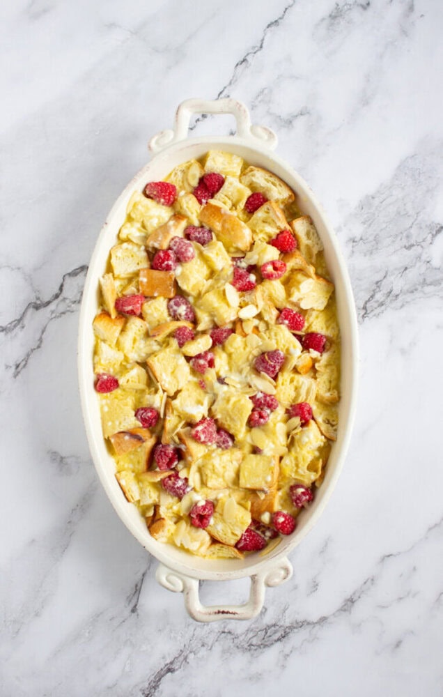 An oval baking dish filled with an unbaked mixture of bread cubes, strawberries, and custard sits on a marble countertop.