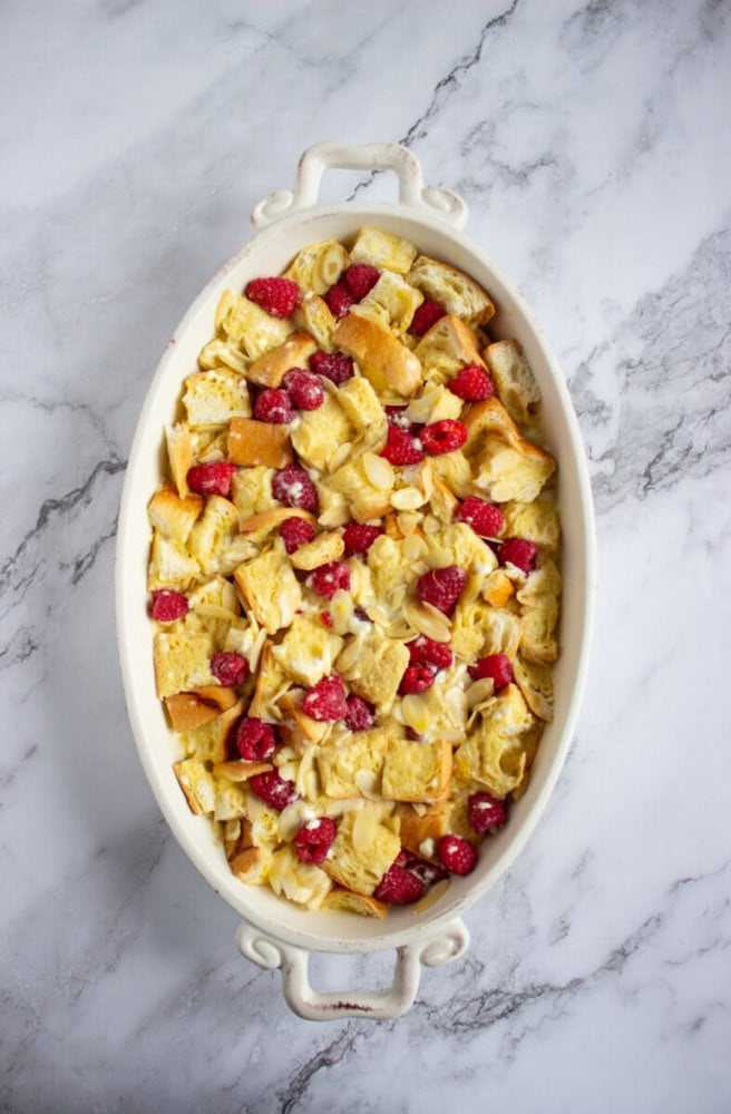 An oval white baking dish filled with bread pudding topped with raspberries and almond slices, set on a marble surface.